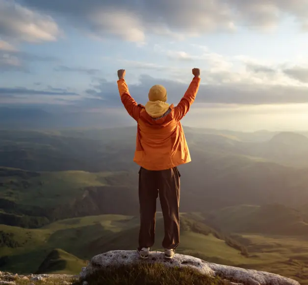 A person in an orange jacket stands triumphantly on a rocky ledge, raising their arms against a scenic backdrop of rolling hills and cloudy skies.