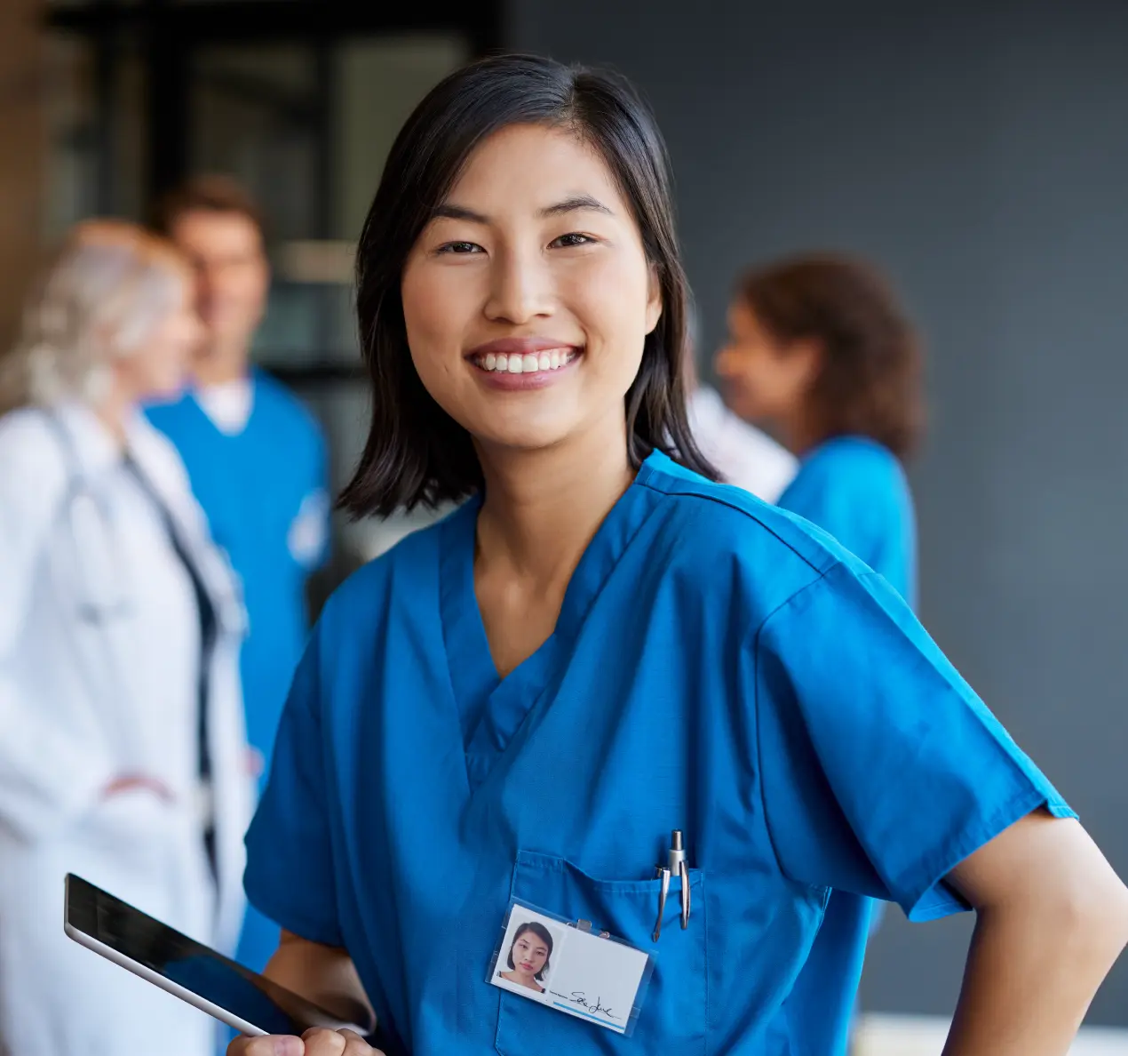 A smiling healthcare professional in scrubs holds a tablet, with colleagues in the background, highlighting a caring medical environment.